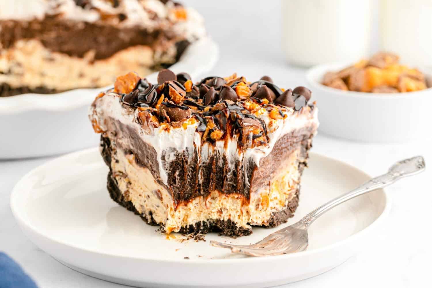 Partially eaten slice of no bake Butterfinger pie showing Oreo crust, peanut butter layer, chocolate pudding, whipped topping, and chopped candy with chocolate chips and syrup.