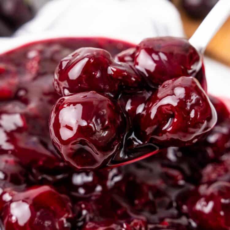 A close-up of a spoon lifting glossy, syrupy whole cherries from a pot of cherry pie filling, showing the thick texture and vibrant red color.