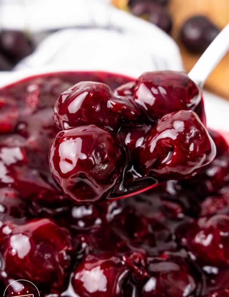 A close-up of a spoon lifting glossy, syrupy whole cherries from a pot of cherry pie filling, showing the thick texture and vibrant red color.
