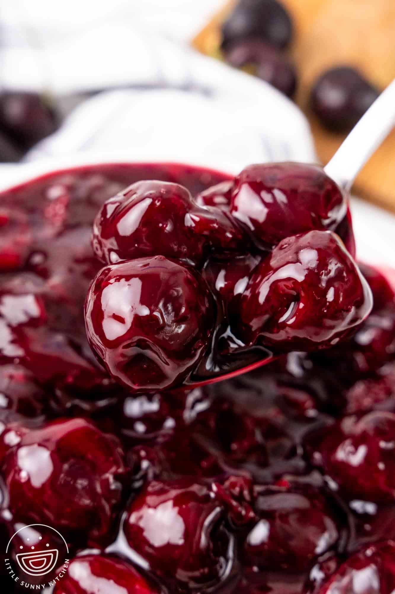 A close-up of a spoon lifting glossy, syrupy whole cherries from a pot of cherry pie filling, showing the thick texture and vibrant red color.