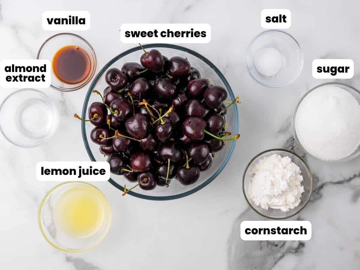 Overhead view of ingredients for homemade cherry pie filling arranged on a marble surface. Includes labeled bowls of sweet cherries, sugar, cornstarch, salt, lemon juice, water, pure vanilla extract, and almond extract.