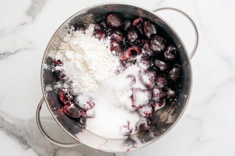 A stainless steel pot with raw pitted cherries, granulated sugar, and a mound of cornstarch before cooking, showing the starting point of the recipe.