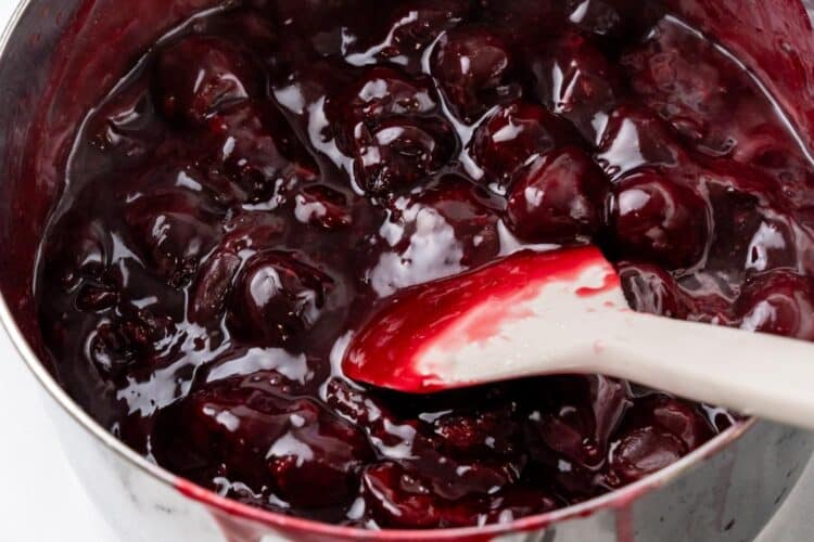 A saucepan with a silicone spatula stirring thickened cherry pie filling. The cherries are coated in a glossy red syrup as they cook down.