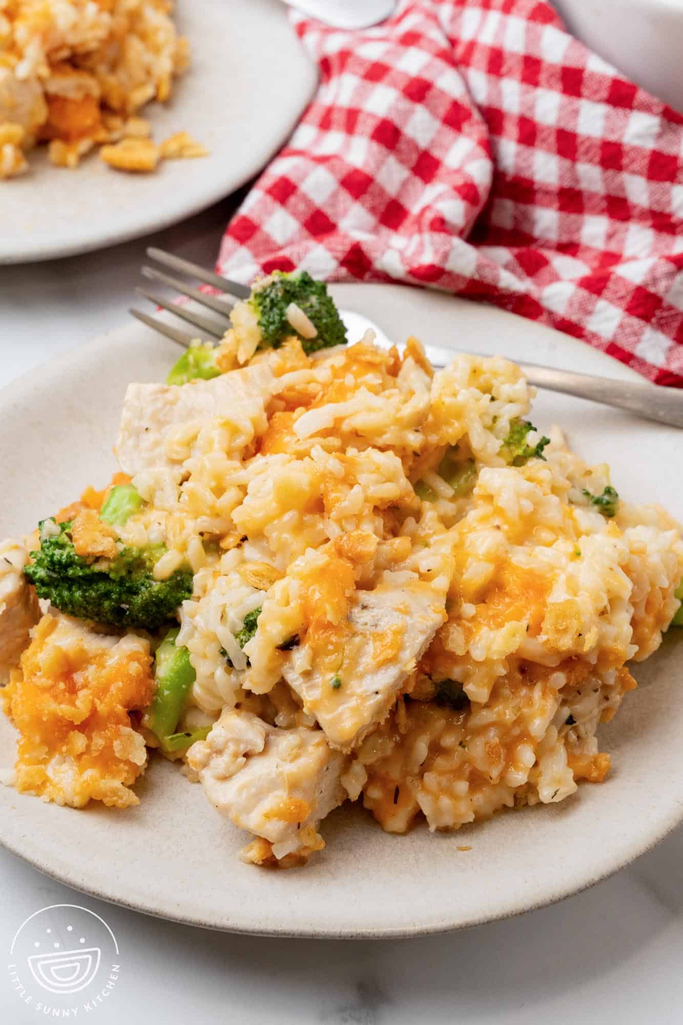 a generous serving of chicken rice broccoli casserole on a plate with a fork. In the background is a red and white checked cloth napkin.