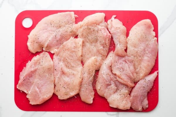 sliced chicken breast cutlets on a red cutting board, seasoned with salt and pepper.