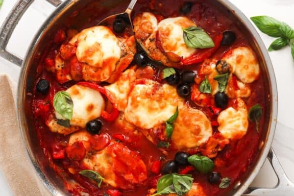 chicken pizzaiola served from a skillet. The chicken is in a tomato sauce made with peppers and mushrooms, topped with cheese, black olives, and basil leaves.