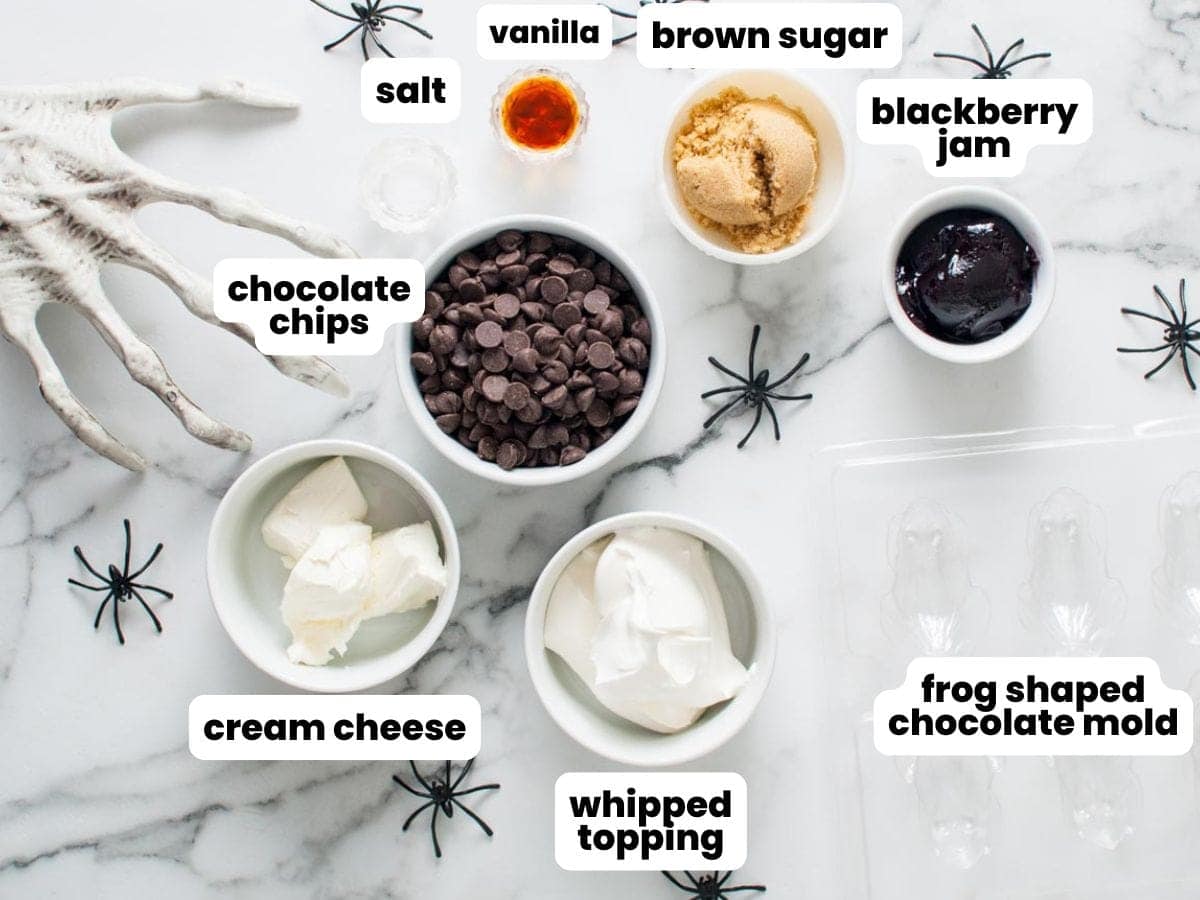 Ingredients for Halloween chocolate frogs laid out on a marble surface, including chocolate chips, jam, cream cheese, whipped topping, and flavorings, surrounded by plastic spiders and a creepy white skeleton hand.