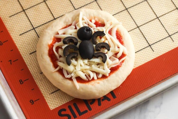 Mini pizza topped with shredded mozzarella and decorated with a black olive spider before baking.