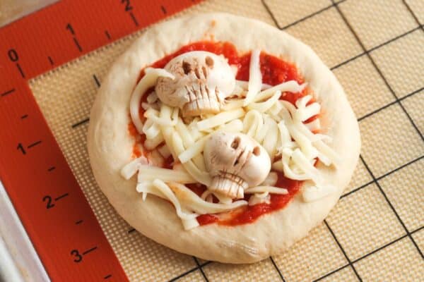 Mini pizza topped with carved mushroom skulls and shredded mozzarella before baking.