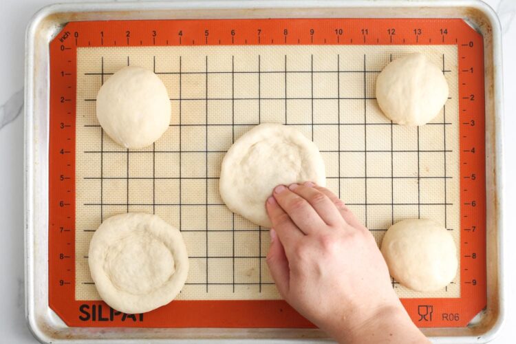 Shaping mini pizza dough portions on a baking sheet lined with a silicone mat. A hand presses down the center to create a crust border.