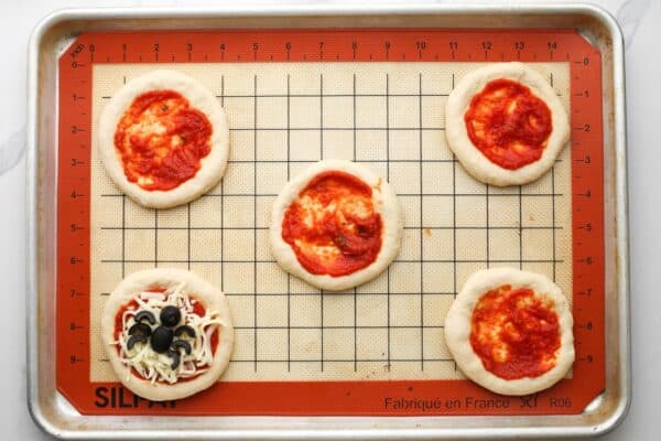 Mini pizza dough rounds with pizza sauce spread on top, arranged on a silicone baking mat.