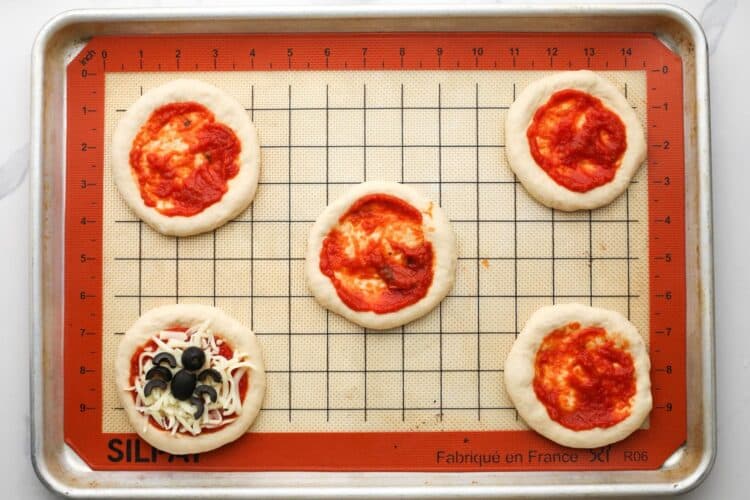 Mini pizza dough rounds with pizza sauce spread on top, arranged on a silicone baking mat.