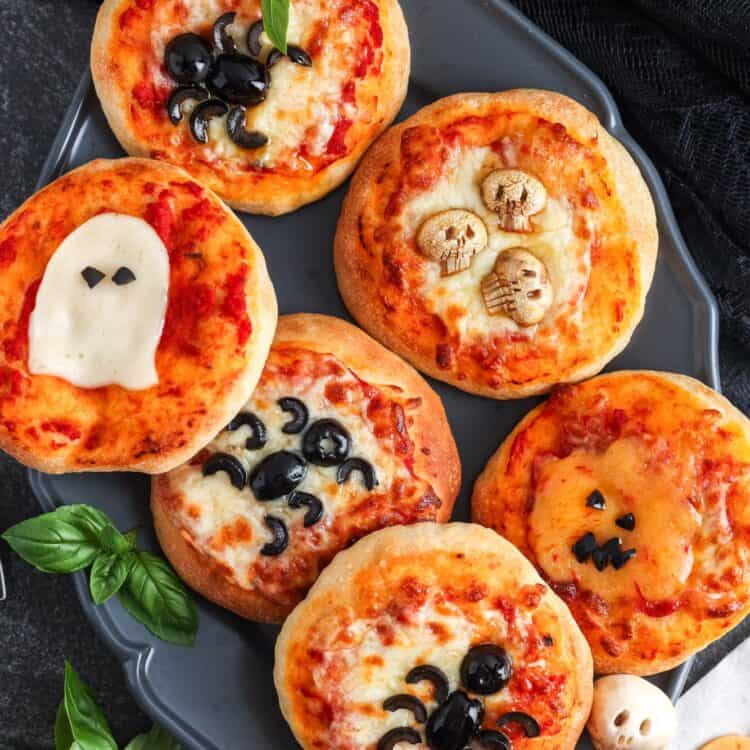 Six mini Halloween pizzas on a dark tray, each decorated with spooky designs like black olive spiders, a mozzarella ghost, a pumpkin face made of cheese, and skull shapes.