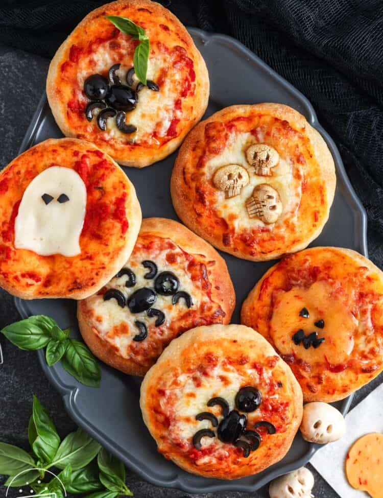 Six mini Halloween pizzas on a dark tray, each decorated with spooky designs like black olive spiders, a mozzarella ghost, a pumpkin face made of cheese, and skull shapes.