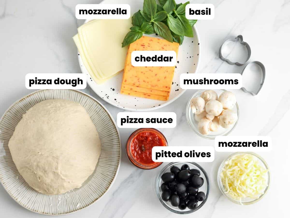 Ingredients for Halloween mini pizzas arranged on a white surface. Includes a ball of pizza dough, a bowl of pizza sauce, pitted black olives, shredded mozzarella, button mushrooms, slices of mozzarella and cheddar cheese, fresh basil, and pumpkin-shaped and ghost cookie cutters.