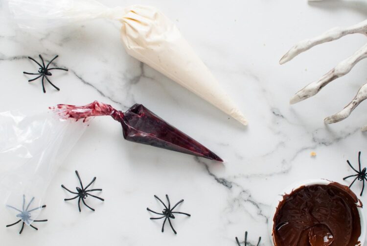 Two piping bags on a marble surface—one with cheesecake filling and one with blackberry jam surrounded by plastic spiders.