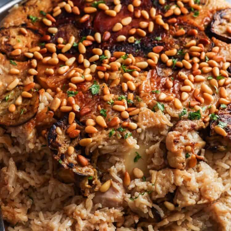 cooked maqluba with chicken and vegetables, flipped over onto a large platter. on top are toasted pine nuts. In the background is a bowl of yogurt and another bowl of cucumber tomato salad.