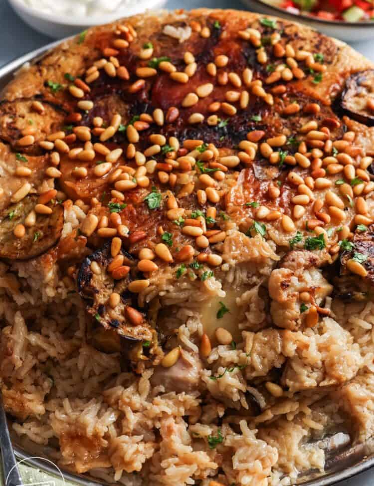cooked maqluba with chicken and vegetables, flipped over onto a large platter. on top are toasted pine nuts. In the background is a bowl of yogurt and another bowl of cucumber tomato salad.