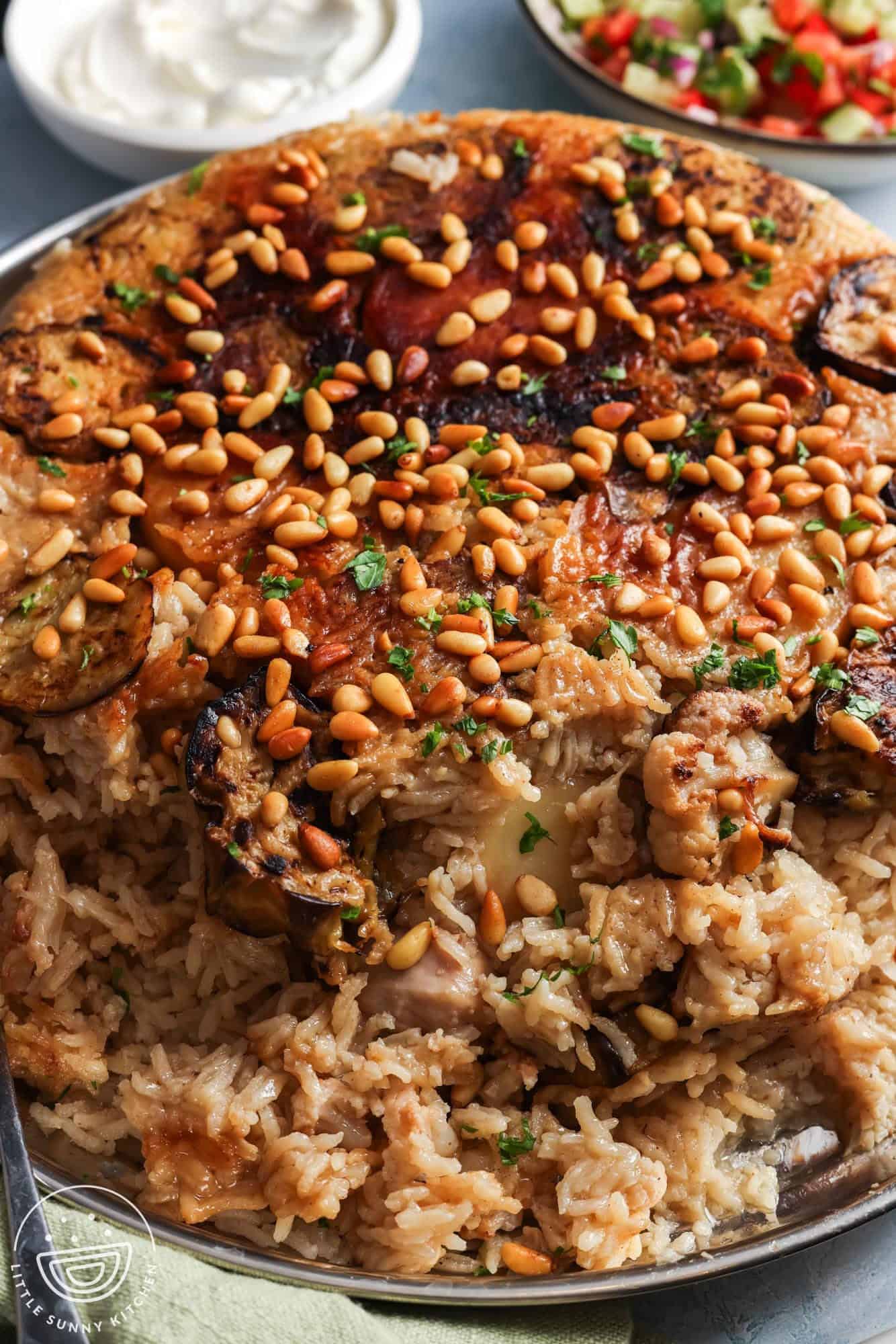 cooked maqluba with chicken and vegetables, flipped over onto a large platter. on top are toasted pine nuts. In the background is a bowl of yogurt and another bowl of cucumber tomato salad.