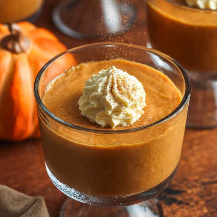 a footed glass dish of pumpkin pudding topped with fresh whipped cream and ground cinnamon