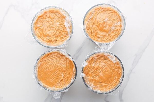 four ramekins of pumpkin pudding. they are covered with plastic wrap that touches the top of the pudding, to avoid a skin forming.
