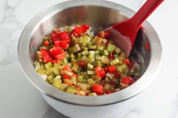 a metal bowl filled with diced strawberries and chopped rhubarb stalks. A red spoon in mixing the berries and rhubarb with flour.