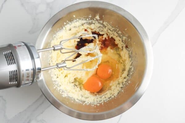a large metal mixing bowl with creamed butter and sugar. Eggs and vanilla have been added and a hand mixer is about to combine them.