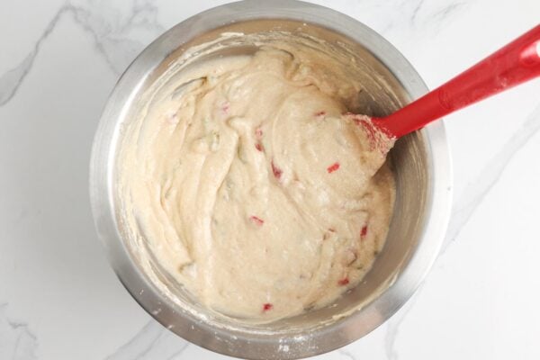 Batter for strawberry and rhubarb muffins in a large metal mixing bowl with a red spoon.