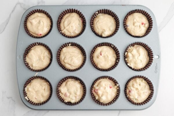 A twelve cup muffin pan lined with brown paper liners that are filled with rhubarb strawberry muffin batter.