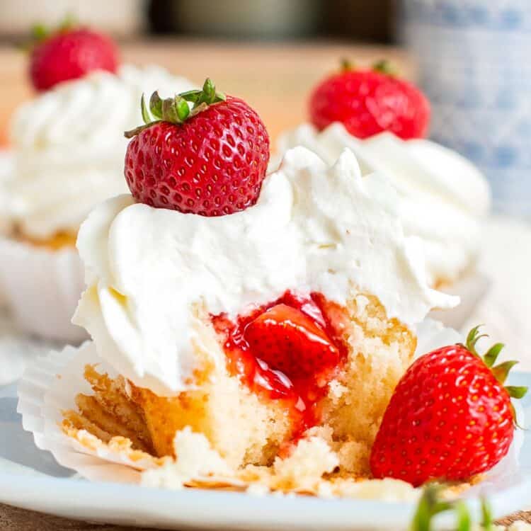 Two strawberry stuffed cupcakes on a plate. Each is topped with fluffy whipped cream and a whole strawberry.