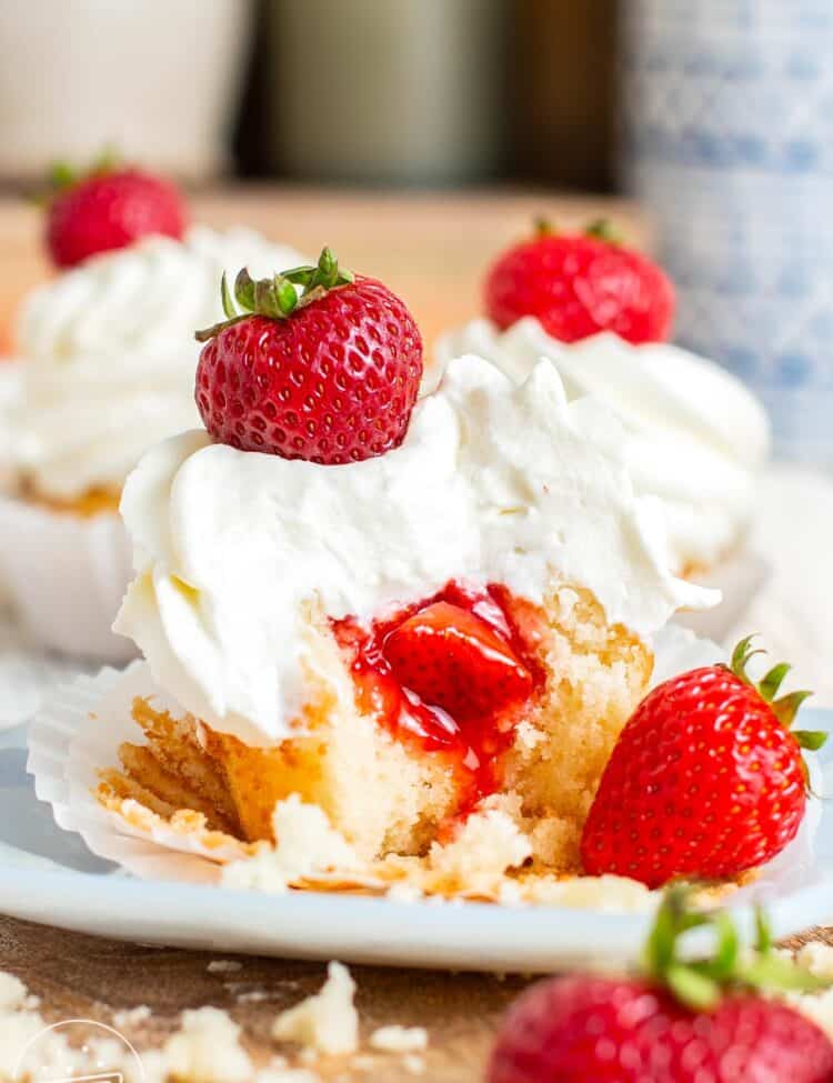 Two strawberry stuffed cupcakes on a plate. Each is topped with fluffy whipped cream and a whole strawberry.