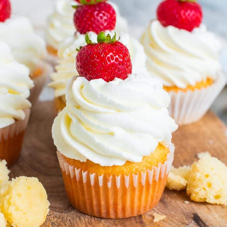 vanilla cupcakes with whipped cream and strawberries lined up on a wooden cutting board.