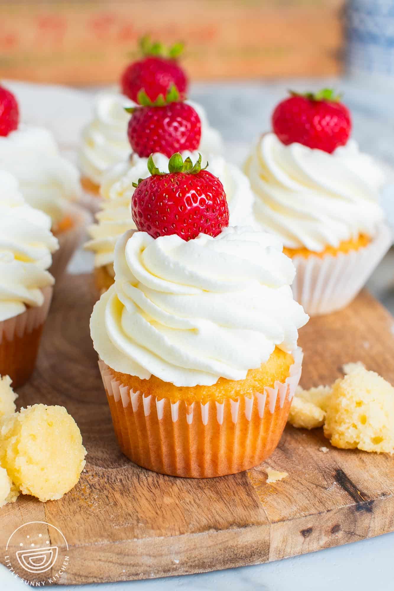 vanilla cupcakes with whipped cream and strawberries lined up on a wooden cutting board.