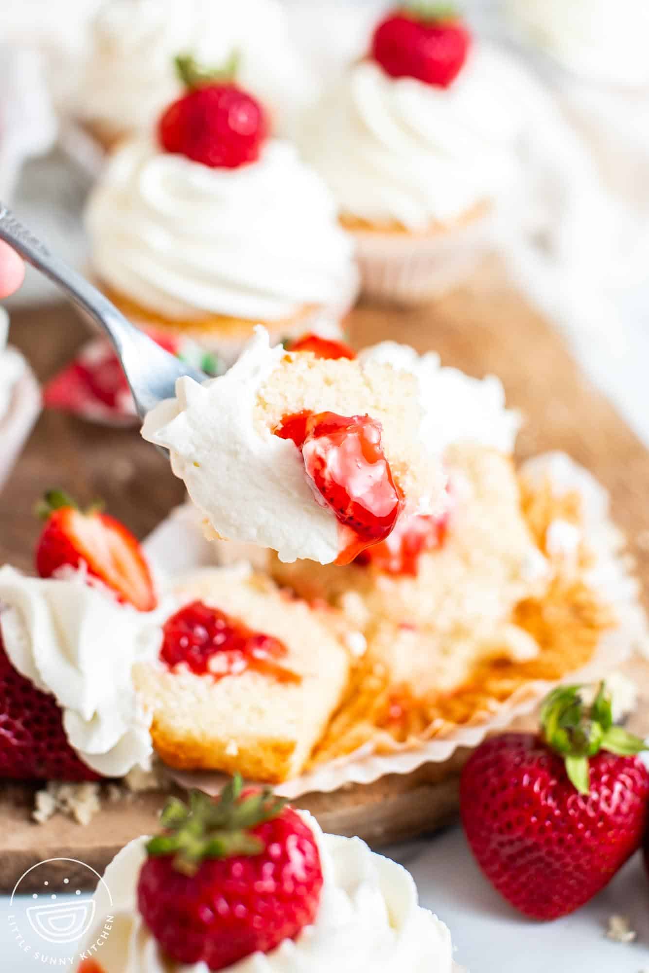 a strawberry shortcake cupcake is cut in half and a fork is taking a bite that shows cake, whipped cream, and strawberry sauce.