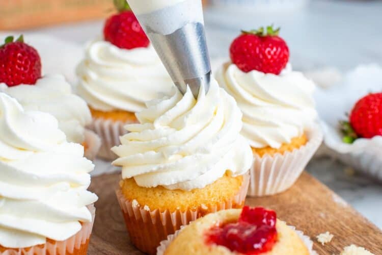 stabilized whipped cream piped onto strawberry filled cupcakes that are set on a wooden board. A whole berry is added to the top of each.