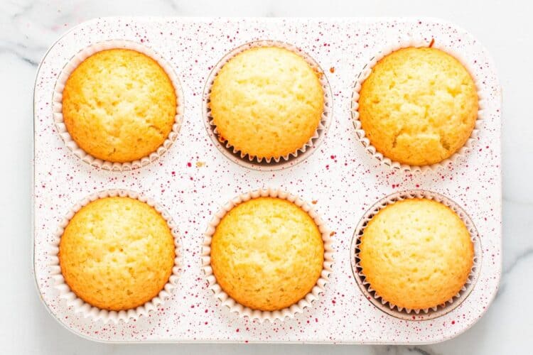 six baked vanilla cupcakes in a white and red speckled cupcake pan.