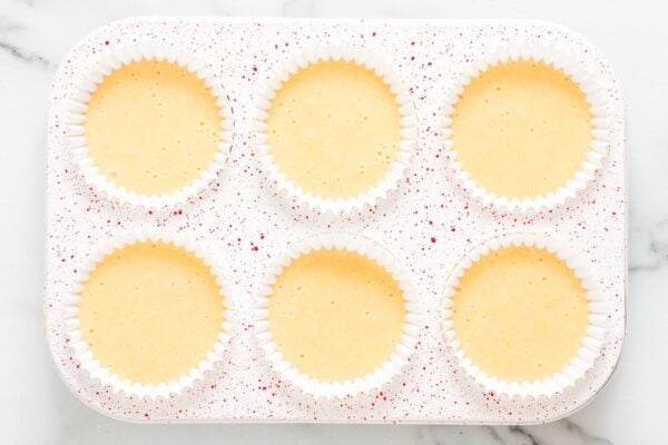 a red speckled 6-well cupcake pan lined with white papers and filled with vanilla cupcake batter.
