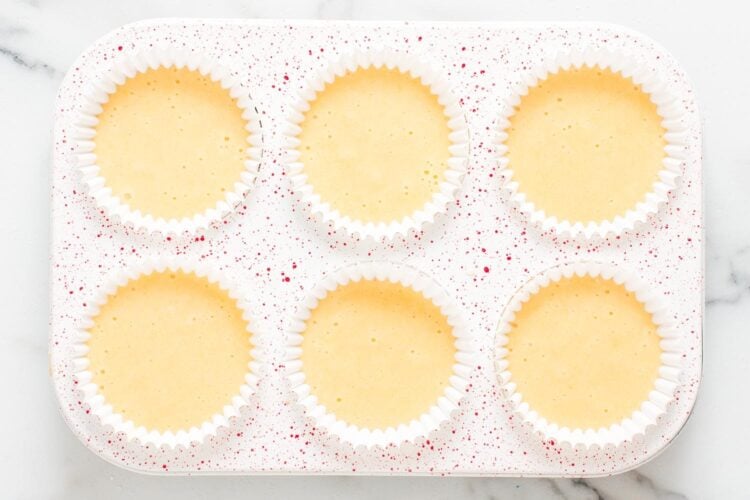 a red speckled 6-well cupcake pan lined with white papers and filled with vanilla cupcake batter.