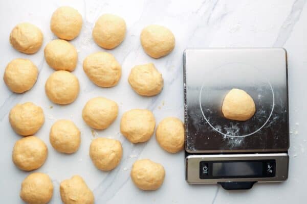 balls of dinner roll dough on the counter. One is being weighed on a stainless steel kitchen scale.