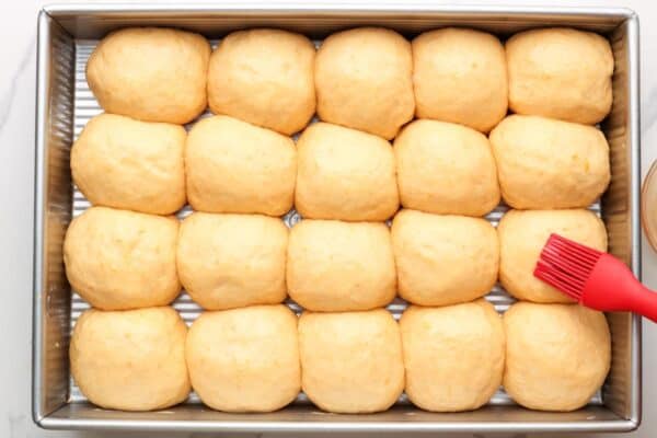 fluffy, risen dinner roll dough with sweet potato in a pan. A red silicone brush is adding milk to the tops before they are baked.