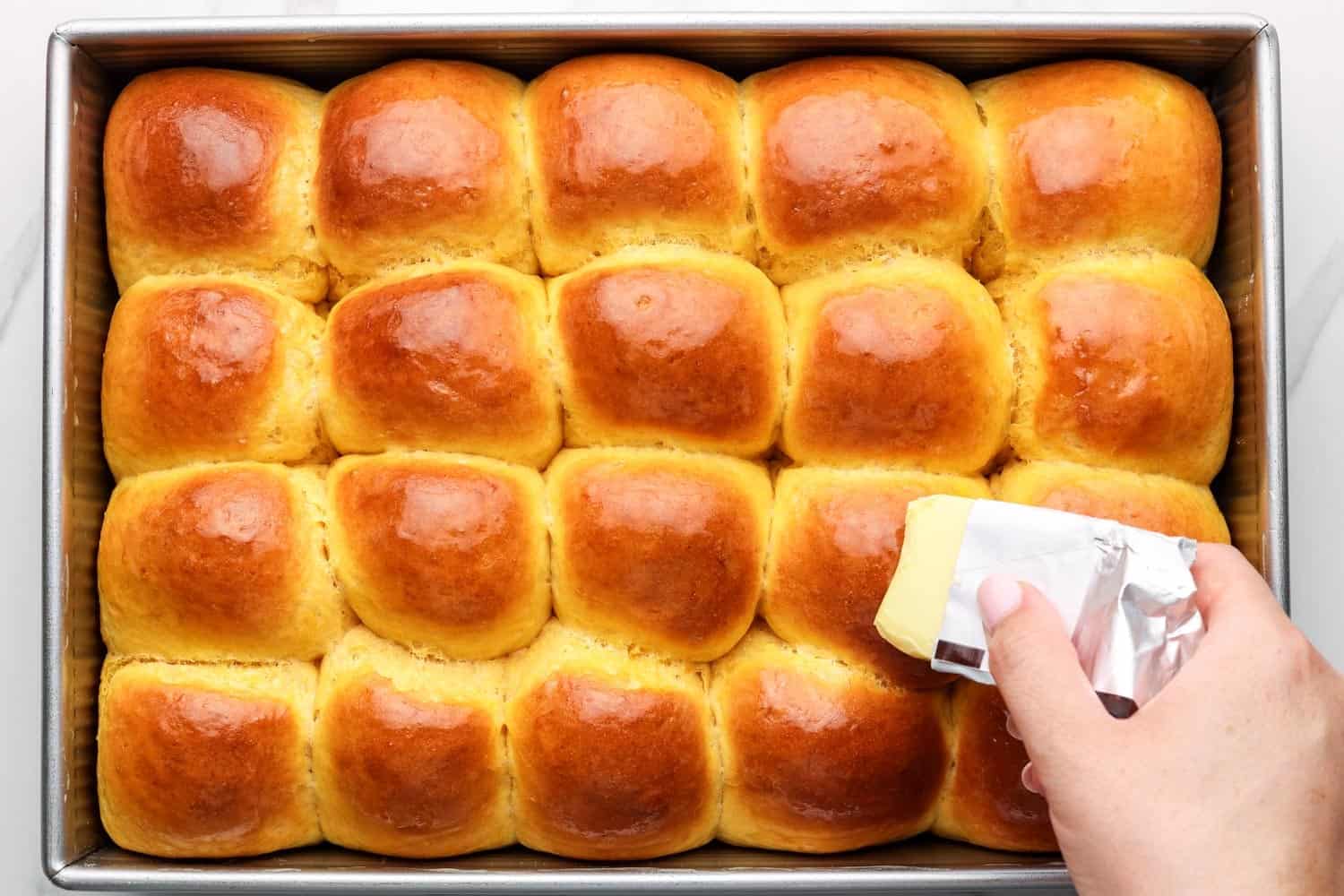 baked sweet potato dinner rolls with browned tops. A hand is adding butter to them while they are still warm.