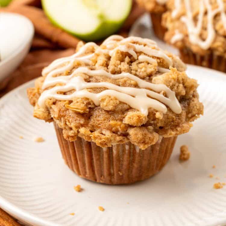 an apple muffin with an oat crumb topping and a drizzle of icing set on a round white plate. In the background are more muffins, green apples, and cinnamon sticks.