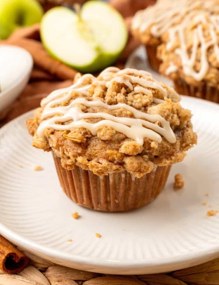 an apple muffin with an oat crumb topping and a drizzle of icing set on a round white plate. In the background are more muffins, green apples, and cinnamon sticks.