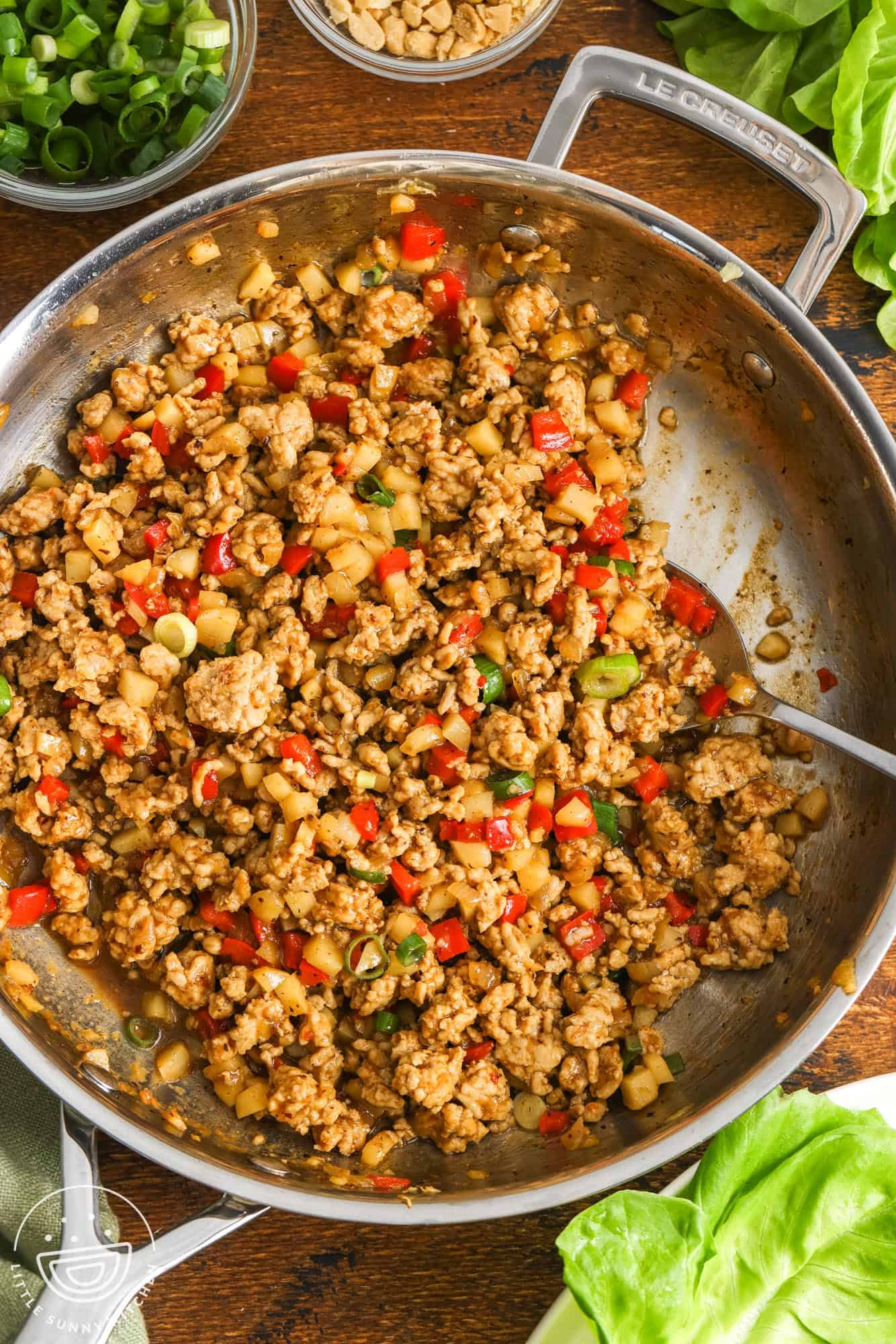 a skillet of asian ground chicken filling on a wooden board. Next to the skillet are butter lettuce leaves for wrapping and small bowls of green onion and chopped peanuts.