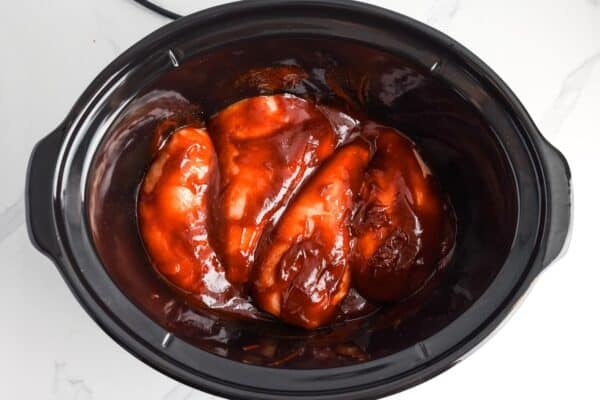 bbq sauce coating four raw chicken breasts in a crockpot.