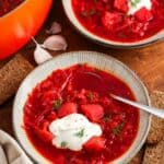two ceramic bowls of deep red borscht soup on a wooden table with slices of dark bread. Each bowl is topped with sour cream and dill.