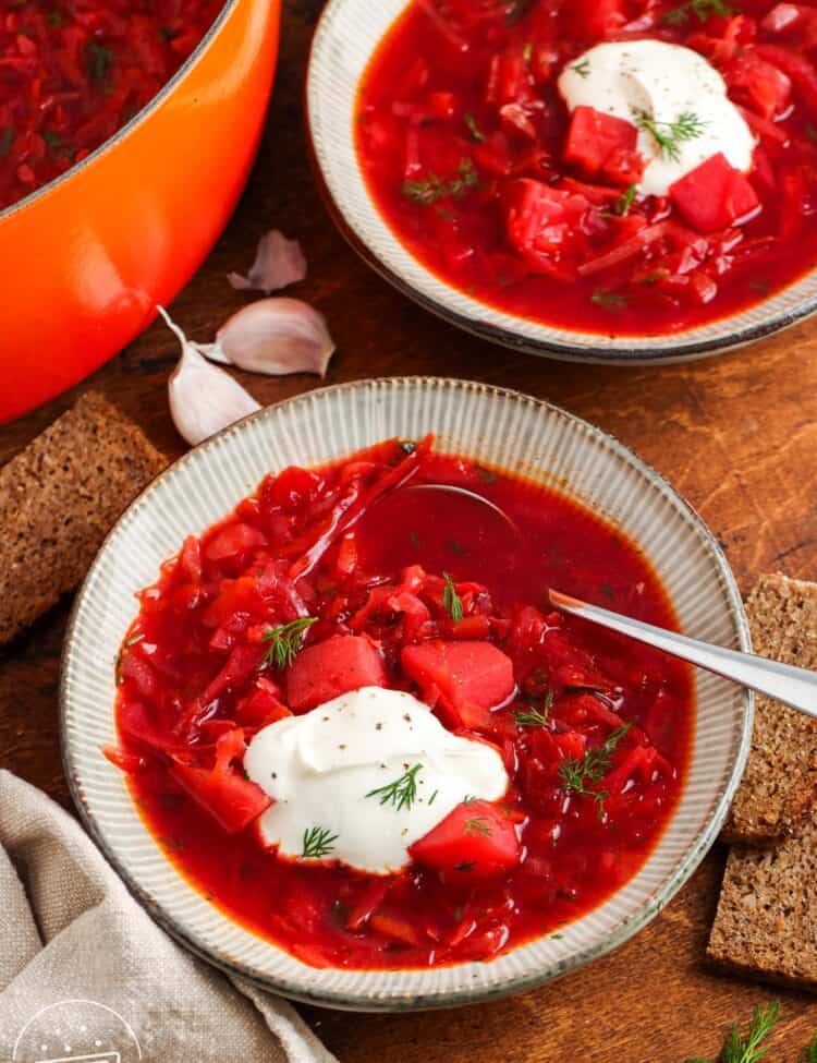 two ceramic bowls of deep red borscht soup on a wooden table with slices of dark bread. Each bowl is topped with sour cream and dill.