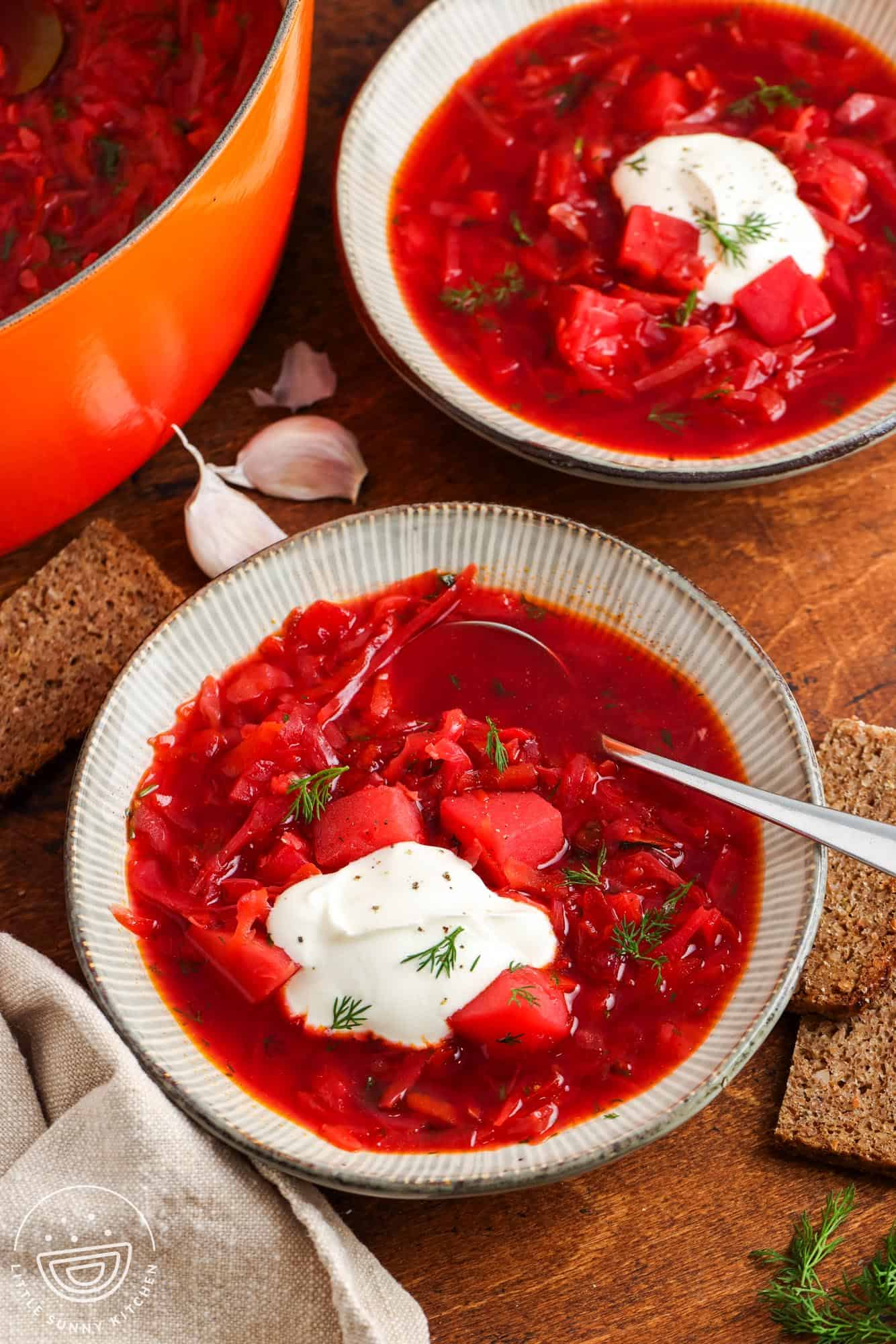 two ceramic bowls of deep red borscht soup on a wooden table with slices of dark bread. Each bowl is topped with sour cream and dill.