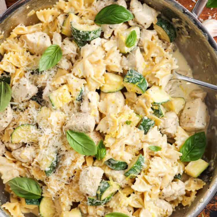 creamy chicken and zucchini pasta served from a stainless steel skillet with a spoon. The pasta is garnished with whole basil leaves and grated parmesan.