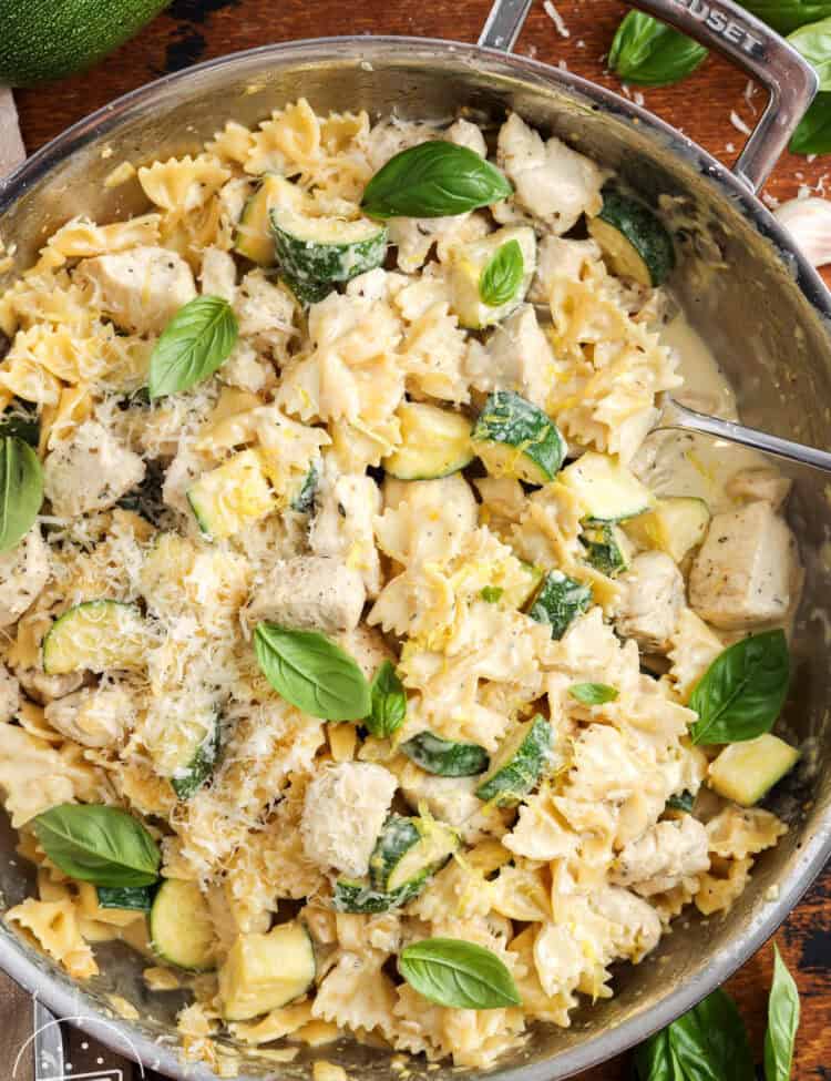 creamy chicken and zucchini pasta served from a stainless steel skillet with a spoon. The pasta is garnished with whole basil leaves and grated parmesan.